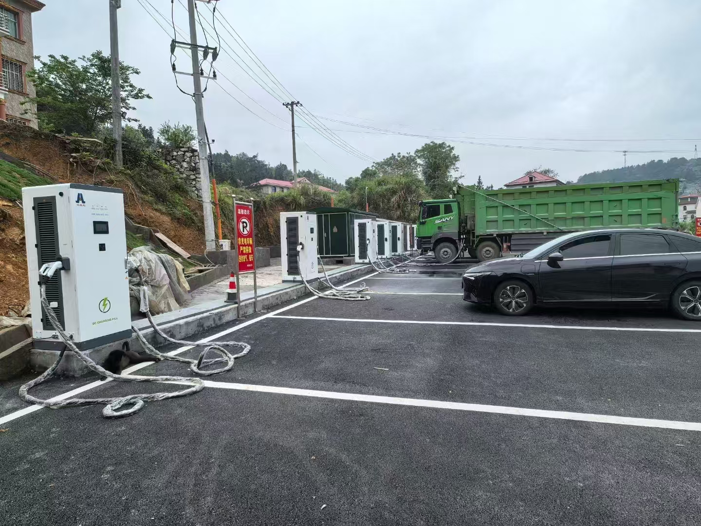 Charging-site overview showing several cabinets, marked bays, a green truck and a passenger vehicle